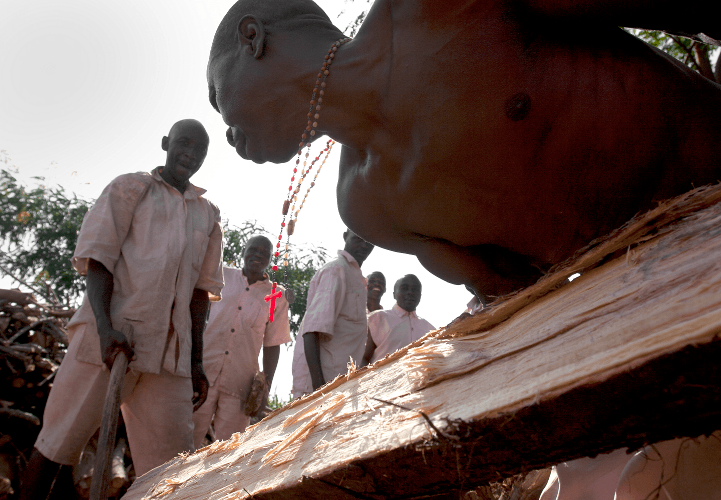 Foto af fanger i et rwandisk fængsel, der under åben himmel er i færd med tungt fysisk arbejde med tømmer.
