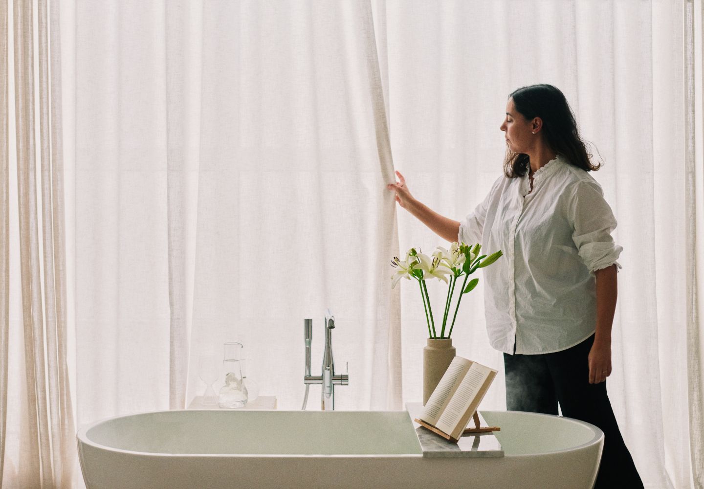 Fulvia Kryger trækker et lyst gardin for bag et badekar, hvorpå der står blomster og et stativ, der kan holde en bog