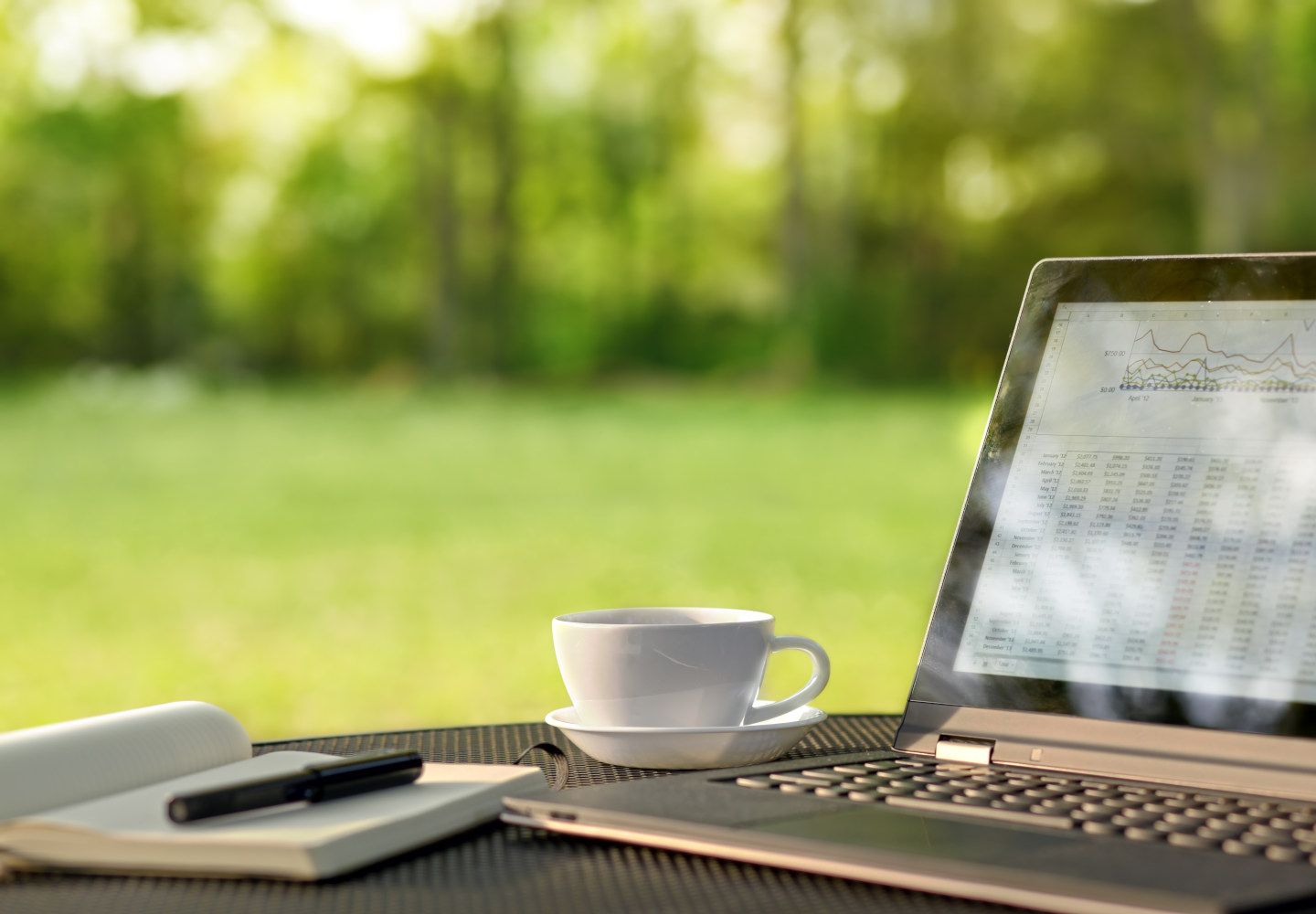 Foto af en laptop, der står på et bord udenfor sammen med en kop og en blok med en pen