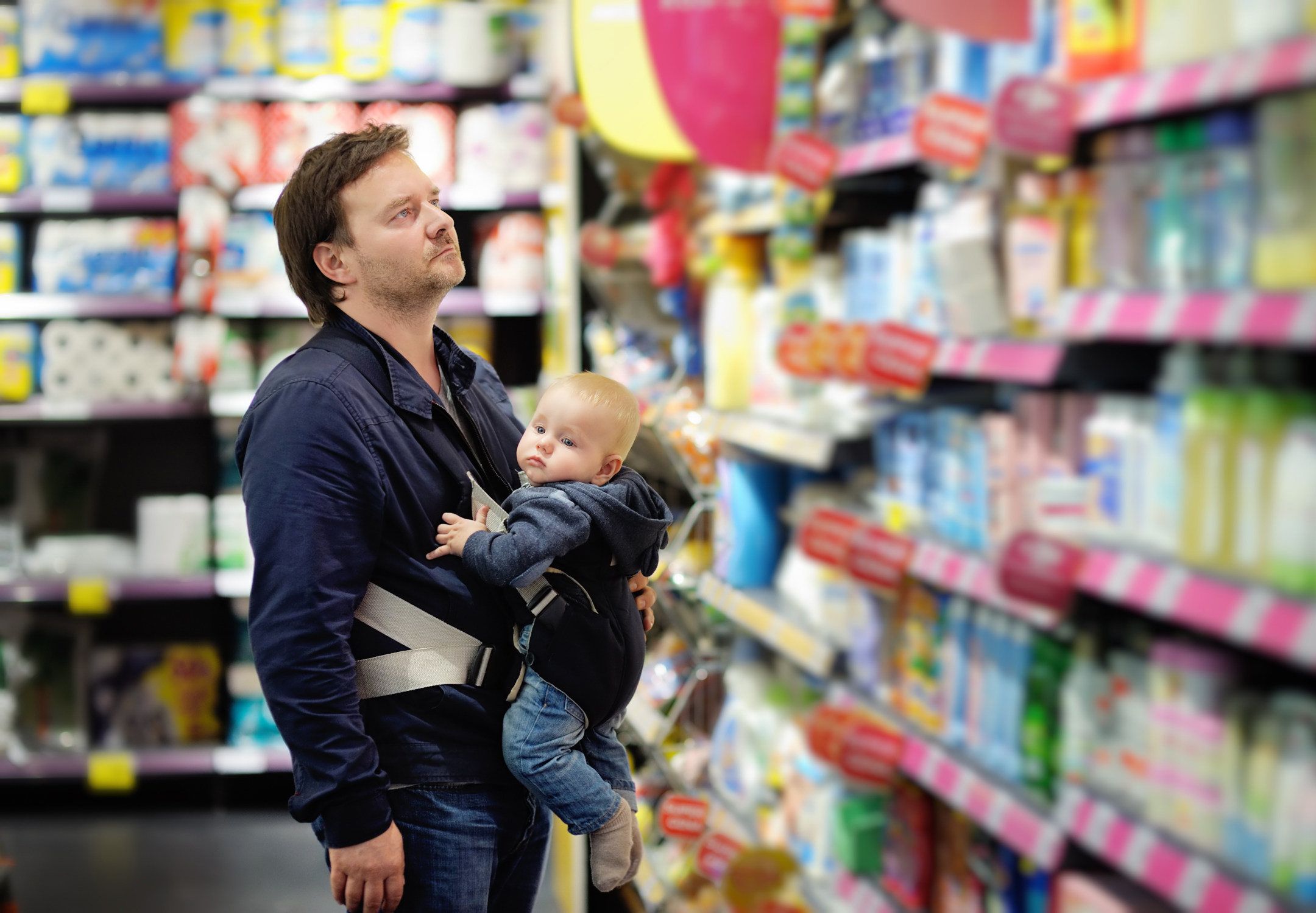 Foto af mand, der står i et supermarked, mens han har et barn i en bæresele på maven