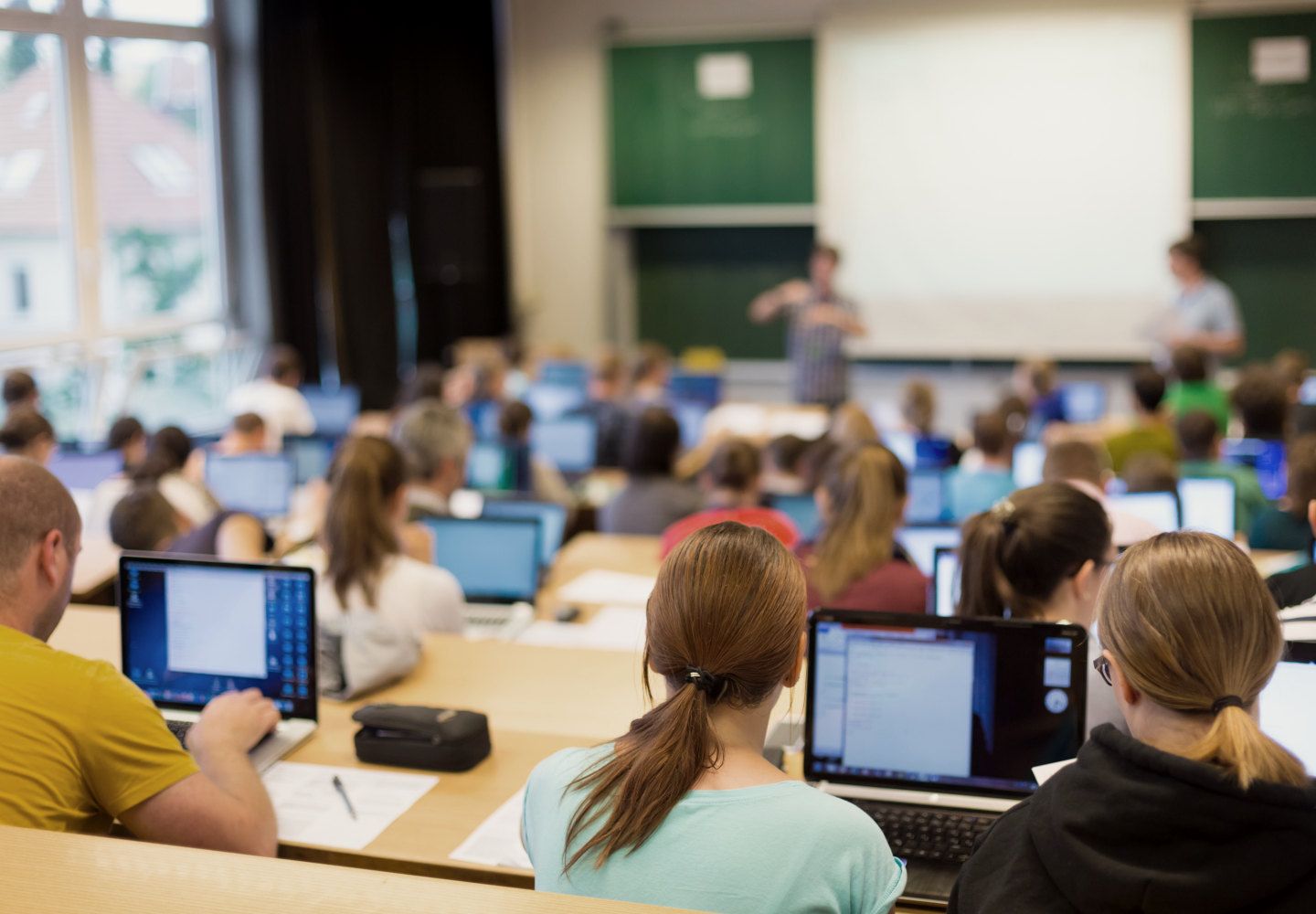 Billede af undervisningslokale med studerende med computere og undervisere ved tavlen